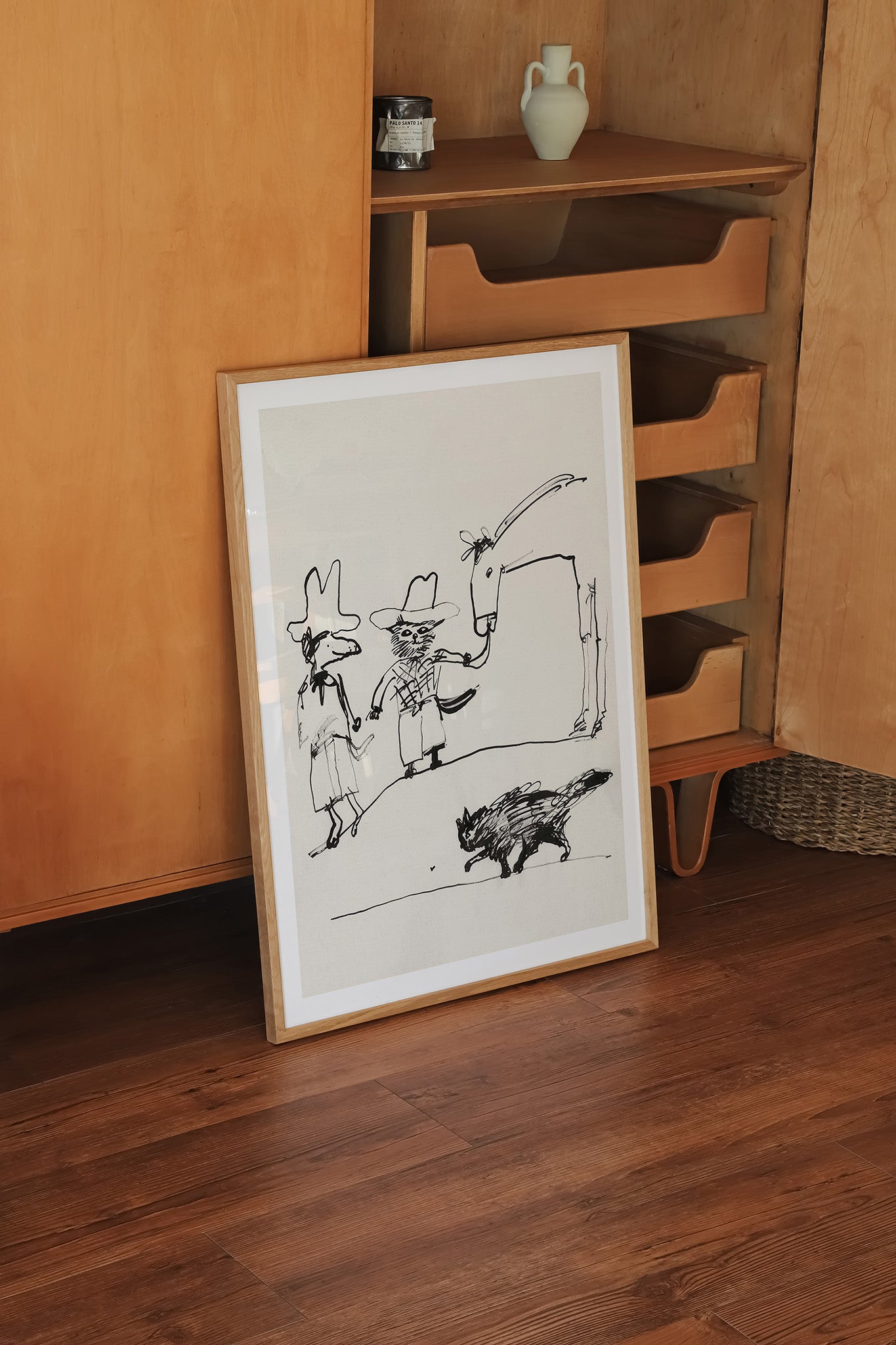 Plakat med enkel sort stregtegning af to figurer med hatte, der står ved en port, mens et lille dyr går forbi i forgrunden. Motivet er indrammet i træramme og står op ad et træskab i et roligt hjemmemiljø.